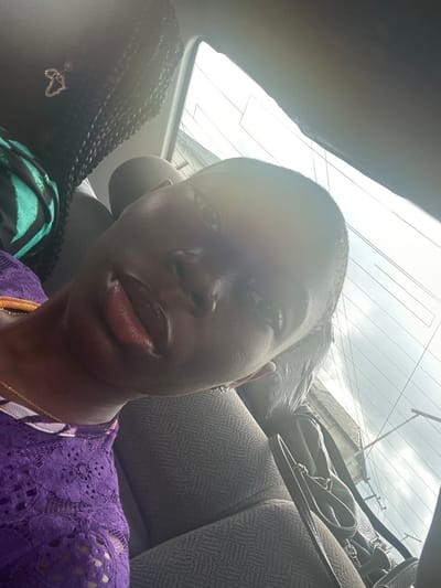The image displays the interior of a vehicle during daylight, featuring two individuals. The primary subject, a person with dark skin, very short dark hair, and full lips, is positioned in the foreground, facing the camera. This individual wears a purple lace top with a patterned trim at the neckline and a thin gold-colored chain. To the upper left, the back of a second individual's head is partially visible, showing dark braided hair and a light-colored, heart-shaped ear or hair ornament. This person wears a green and black striped garment. The vehicle's interior includes gray, fabric-covered seats with a subtle patterned texture. A dark handbag or satchel rests on the seat to the right of the primary subject. Through the rear window, a bright, overcast sky is visible, with faint, indistinct structures or power lines in the distance. The scene is located within Rumuogonoma, Nigeria.
