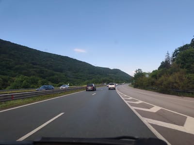 The image depicts a highway scene with a mountain covered in dense green foliage to the left and a line of trees to the right. The sky is a clear, pale blue, suggesting it might be early morning or late evening. Several cars are visible on the highway, including a dark SUV in the foreground, a white pickup truck further ahead, and other vehicles in the distance. The road has white lane markings, and a metal guardrail runs along the left side of the highway, separating it from the mountain. In the distance, power lines are visible on the crest of the mountain. The scene appears to be located on a highway in or near Potop, Bulgaria.