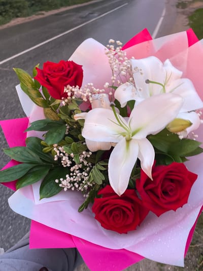 A close-up, overhead shot showcases a vibrant bouquet of flowers arranged in a pink and white paper wrapper. The bouquet is dominated by three deep red roses, their petals velvety and tightly furled. Interspersed are elegant white lilies, their large, trumpet-shaped blooms unfurling gracefully. Delicate sprigs of baby's breath, with their tiny white flowers, add a touch of airiness to the arrangement. Lush green foliage forms a verdant backdrop for the prominent blossoms. The bouquet appears to be damp, with small water droplets visible on the petals and wrapping paper, suggesting recent rain or mist. The background is blurred, revealing a grey asphalt road with a white stripe running down the center, hinting at an outdoor setting, possibly near a roadside. A hint of greenery borders the road. The scene is bathed in natural light, though the specific time of day is not clearly discernible. The overall impression is one of natural beauty and thoughtful presentation. There is no visible text in the image.
