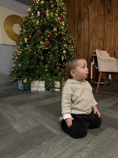 An indoor scene in Kalinovo, Russia, features a young child kneeling on a dark gray herringbone-patterned floor. The child, a toddler with light brown hair, wears a horizontally striped hooded sweatshirt, a white long-sleeved undershirt, and dark corduroy pants. The child faces right, looking off-camera with a slightly open mouth. In the background, to the left, stands a large, illuminated artificial Christmas tree decorated with warm white lights and red, gold, and blue ornaments. Several wrapped presents, including blue with snowflakes, white with polka dots, and green with a gold ribbon, are visible beneath the tree. The wall behind the tree on the left is dark gray, featuring a large, golden circular art piece on a white canvas. The wall behind the child and to the right is composed of vertical wooden planks. Partially visible on the far right is a light-colored upholstered chair and a child's high chair frame.