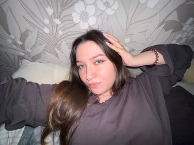 The image shows a young woman lying down with her head on a pillow. She is wearing a dark grey t-shirt and has long, dark brown hair. Her right hand is resting on her head, and she is wearing a beaded bracelet on her left wrist. The background appears to be wallpaper with a floral pattern. The scene is indoors.