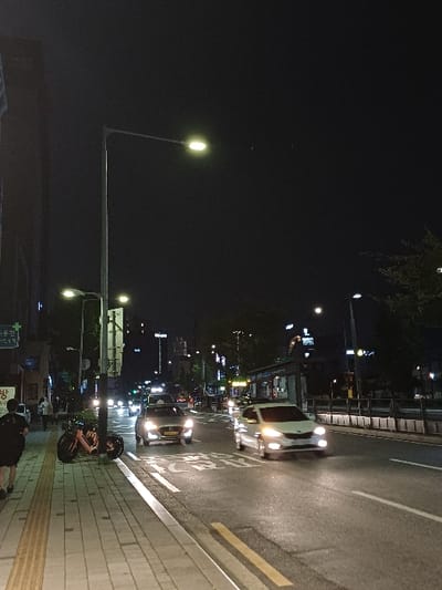 The image depicts an urban street scene at night in 서대문구, South Korea. The perspective is from a sidewalk looking down a multi-lane road.
On the left, a paved sidewalk with light grey square tiles and a strip of yellow tactile paving is visible. A single individual, appearing to be male and dressed in dark clothing (dark t-shirt, dark shorts), walks away from the camera on the sidewalk's left edge. Immediately to the right of this individual, a dark-colored scooter or small motorcycle with orange accents is parked upright next to a street light pole. Behind the individual and scooter, a building facade with illuminated signs bearing Korean characters, including "방" and partially "대문구", is present.
In the center-midground, a wide asphalt road extends into the distance. White lane markings and a partial white pedestrian crossing symbol are painted on the asphalt. Two vehicles are prominently positioned in the road's foreground lanes, both with active headlights, moving away from the viewer. The vehicle in the left lane is a light-colored sedan; the vehicle in the right lane is a light-colored compact car or small SUV. Numerous additional vehicles are visible further down the road, indicated by their illuminated headlights and taillights. The road surface appears damp, reflecting artificial light.
Along the right side of the road, a concrete barrier separates the main thoroughfare from a paved area where a bus stop shelter structure is visible. Sparse trees or shrubs line this area and extend into the background.
Overhead, the sky is dark, characteristic of nighttime. Multiple street lights with bright, yellowish illumination are positioned along the road, providing the primary light source in conjunction with vehicle head and tail lights. Distant buildings with illuminated windows are visible on both sides of the street in the background.