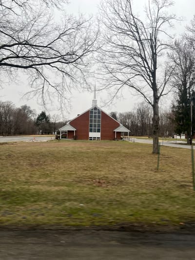 This image presents a brick church building situated in a large, discolored grassy field in the Town of Fishkill, United States of America. The church features a prominent white steeple and a tall, multi-paned vertical window arrangement on its red brick facade, which is accented with white trim. Covered porch-like extensions with white railings flank the main entrance, with one side including an access ramp. The grassy field surrounding the church shows a mix of brown and green, indicative of a dormant season. To the left of the church, a paved area, possibly a parking lot, contains at least two orange traffic cones. A paved driveway or path curves away to the right of the building, leading towards a small white structure further in the distance. The background consists of dense lines of bare deciduous trees and some evergreen conifers. The sky is uniformly overcast and light grey. No people or active interactions are visible in the scene.