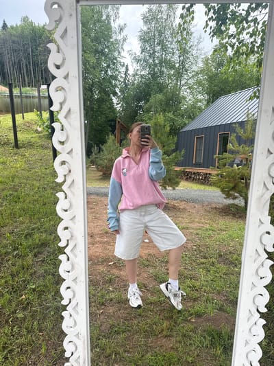 The media file depicts a mirrored reflection of a person standing outdoors in what appears to be a rural setting. The person is wearing a pink and light blue hooded sweatshirt with a Hello Kitty graphic on the front, white shorts, white socks, and white athletic shoes. They are holding a smartphone in their right hand, taking the photograph.

In the reflection, a dark-colored building with a sloped roof and rectangular windows is visible behind the person. There is a stack of firewood next to the building and some pine trees. The ground is covered in grass and dirt. The sky is overcast, suggesting a cloudy day. In the distance, a body of water and more trees are visible. The overall scene conveys a casual, outdoor atmosphere. The white decorative trim of the mirror frame is prominent in the foreground. No audible dialogue or explicit emotions are discernible from the static image. The location is identified as Novaya Vozhoika, Russia.