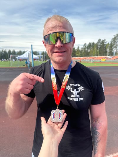 A middle-aged man is standing on a track and field, wearing sunglasses, a black t-shirt with Russian text, and a medal around his neck with “Russia” printed on the lanyard. He is pointing to the medal held by a hand. The background includes stadium seating, grass, trees, and a blue sky with clouds. The shirt says "KРОССФИТ C.F.M.P.Z. ПОЛЯРНЫЕ ЗОРИ". This translates to "CrossFit S.F.M.P.Z. POLAR ZORI". Polarnie Zori is a city in Russia.