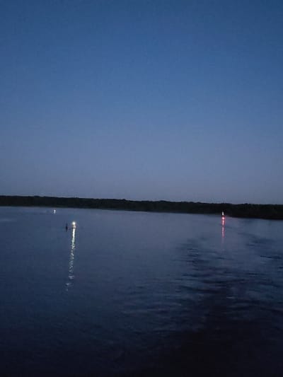 The image depicts a dark, expansive body of water at dusk, under a clear, deep blue sky. A thin, dark line of trees or vegetation runs along the horizon. Two small, illuminated markers are visible on the water. One marker, appearing to be a buoy, emits a bright white light and its reflection shimmers on the water's surface. Another marker, further to the left, is a smaller point of light, possibly red or orange, also with a faint reflection. The water is calm, with gentle ripples catching the faint light. The overall scene suggests a quiet, possibly remote location, devoid of human activity except for the distant markers. The image is believed to have been taken in Irdomatka, Russia, a location known for its natural landscapes.