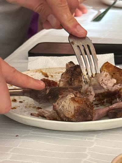 A close-up shot focuses on a person's hands using a fork to pull apart pieces of cooked meat on a white plate. The meat appears to be a roast, possibly pork or beef, with visible layers of muscle and fat. Some pieces are darker brown, indicating they are well-cooked or have a crust, while others are lighter pinkish-brown. A dark red sauce or condiment is visible on the plate near the meat. The plate rests on a white tablecloth with a subtle pattern. In the background, another fork with a dark handle is partially visible, as is a portion of a dark rectangular object, possibly a phone or tablet, lying flat. The overall scene suggests a meal is being prepared or consumed. The image does not provide any information that would allow for its location to be identified in Pavlohrad, Ukraine, or any other city.