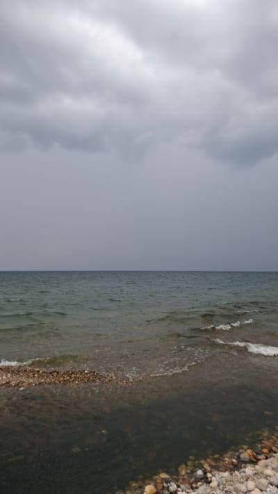 The image depicts a gloomy, overcast day at a stony beach. The sky is filled with heavy, gray clouds, obscuring any hint of sunshine. Below the sky, a vast expanse of dark, choppy water stretches to the horizon, with gentle waves lapping at the shore. The beach itself is covered in a mixture of small and large stones, some wet and dark from the water, others dry and lighter in color. The overall impression is one of a somber, natural landscape, possibly at the edge of a large body of water like Lake Baikal, given the location specified in Priboi, Russia. There are no people or man-made structures visible in the frame.