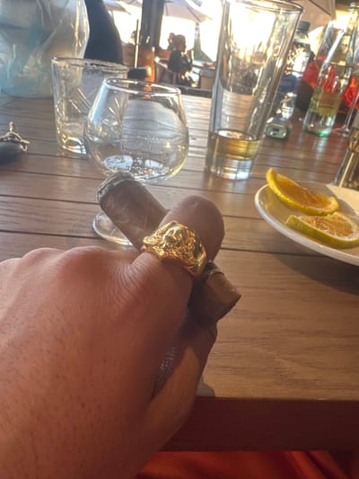 A close-up shows a hand holding a lit cigar, adorned with a large gold skull ring. A wooden table with several glasses is visible in the background. A small plate of orange slices sits at the top of the image. The scene is brightly lit, casting strong shadows. The image has a slight tilt.