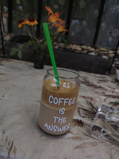 The image shows a glass with coffee and a straw, placed on a table with a patterned tablecloth. The glass has white text that reads "COFFEE IS THE ANSWER". In the background, there are orange flowers and greenery, suggesting an outdoor setting. A glass ashtray is also visible on the table. The scene appears to be in Каменар, Bulgaria, possibly a cafe or a garden setting. There are no people in the image.