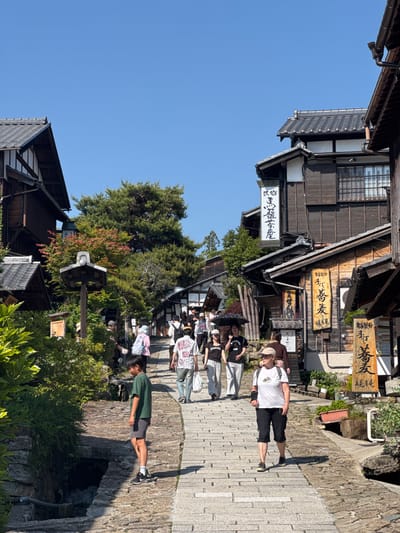 This image shows a street in the city of Nakatsugawa, Japan. The street is paved with cobblestones and lined with traditional Japanese buildings. There are several people walking along the street, including one man wearing a white t-shirt and black shorts and a person wearing a green t-shirt and grey shorts. The buildings are made of wood and have traditional Japanese roofs. There are also some trees and plants along the street. The sky is blue and clear.
