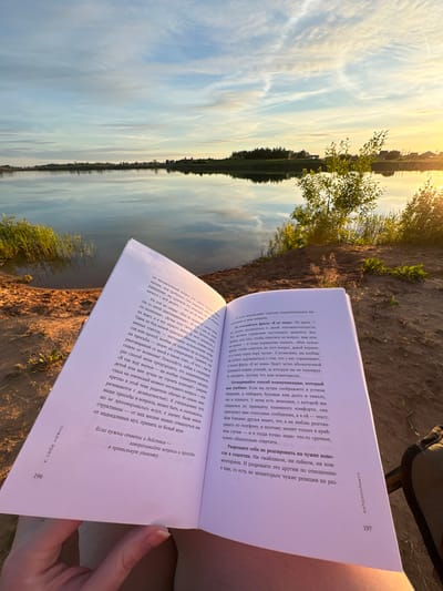 A book is open and held by an unseen person, its pages are illuminated by the golden light of a sunset. The book's pages are filled with Russian text. The book is set against a backdrop of a tranquil lake, its surface reflecting the soft colors of the sky. Lush green grass and trees frame the scene, with the silhouette of a distant treeline visible on the opposite bank. The overall atmosphere is serene and peaceful, suggesting a moment of quiet contemplation in nature. The location is identified as Sapunov Bor, Russia.