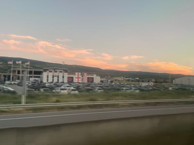 In Gorovani, Georgia, a wide shot captures a commercial building with a large parking lot filled with numerous vehicles. The building features prominent flags, including the Georgian flag, along with what appear to be United States and United Kingdom flags, displayed on poles above the structure. The building's facade includes the Georgian flag and the Union Jack as decorative elements. The sky above is a pale blue transitioning into soft pink and orange hues, indicating either dawn or dusk, with scattered clouds. A highway is visible in the foreground, separated from the parking lot by a grassy median with a guardrail. The background consists of rolling hills and a distant mountain range under the soft light of the sky. The overall scene depicts a commercial establishment in a semi-rural setting.