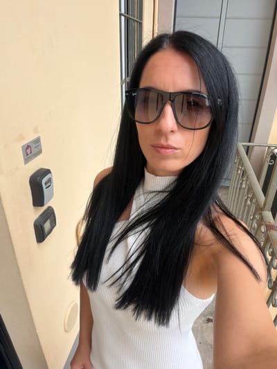 A medium shot captures a woman with long, dark hair and sunglasses, wearing a white, ribbed, sleeveless top. She is standing outdoors in front of a light beige wall, possibly the exterior of a building. Mounted on the wall are two grey boxes, one above the other, and a sticker with a house logo and text that reads "Alessia's Flat." To the right of the woman, a metal railing with decorative elements is visible, suggesting a balcony or staircase. The background consists of a building facade. The scene appears to be a residential area, with the presence of the key safe and building signage pointing to Buccinasco, Italy, as the location.