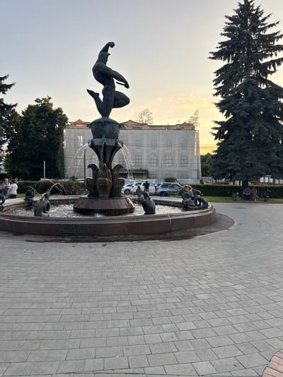 This image depicts an outdoor scene centered around a fountain. The fountain features intricate bronze sculptures, including a figure in mid-air holding a dagger, and various mythological creatures such as sea serpents. Water is actively spraying from the fountain, creating a dynamic element. The fountain is situated in a paved area, likely a public square or park, with a stone walkway bordering it. In the background, trees with lush green foliage are visible, along with a building that appears to be under construction or renovation, indicated by scaffolding. A few people are present in the scene, some seated on a bench near the trees and others near the fountain. The time of day appears to be late afternoon or early evening, with the warm glow of the setting sun illuminating the sky and casting soft light on the surroundings. The weather seems clear and pleasant. There is some visible text on the base of the fountain, which appears to be in Cyrillic script. The overall atmosphere is one of a tranquil urban public space.