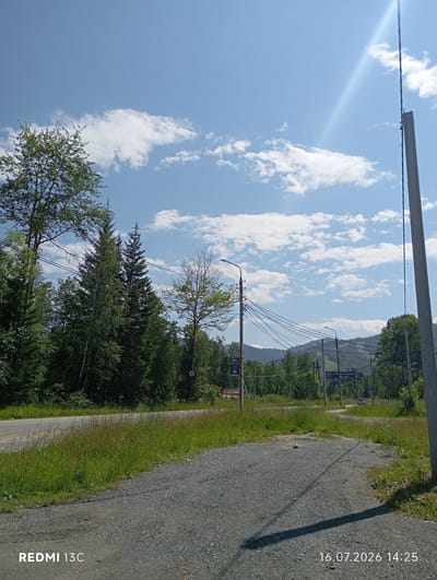 The image captures a sunny day in Baykalsk, Russia, with a clear blue sky dotted with white clouds. In the foreground, a gravelly path leads towards a paved road. Lush green grass and trees line the side of the road, with a dense forest in the background. Mountains are visible in the distance. Several utility poles with electrical wires are present, suggesting infrastructure in the area. A few signs are visible along the road, but their text is not legible. The overall scene depicts a natural, rural setting with some signs of human development.