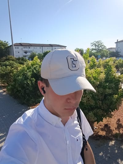 A man wearing a white cap with a stylized letter "C", a white button-down shirt, and possibly a backpack, is standing outdoors near a bush with light yellow flowers and a building in El Port De Sagunt, Spain. He has a wireless earbud in his left ear. The location seems to be a street with a pedestrian crossing sign visible. The ground is partially covered with light brown gravel or small stones. The sky is clear and blue.