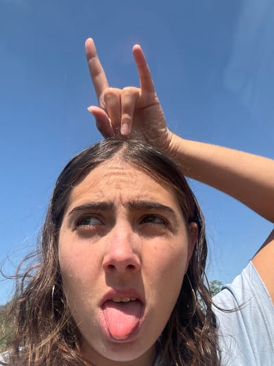 A close-up, low-angle shot depicts a person outdoors in Or Yehuda, Israel, under a clear, bright blue sky. The subject, with dark, wavy hair parted in the center, has their head tilted slightly upwards, eyes looking towards the upper right. Their tongue is extended from an open mouth, and their eyebrows are slightly furrowed. A silver hoop earring is visible on the subject's left ear. The subject's left hand is raised directly above their head, positioned at the hairline, forming the "sign of the horns" gesture with the index and pinky fingers extended. A light-colored garment, possibly blue or grey, is visible on the subject's right shoulder and arm.