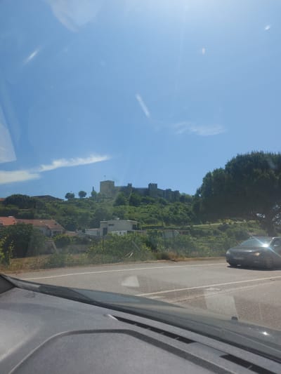This image presents a bright, sunny daytime view from the passenger side of a moving vehicle, looking out at a scenic landscape featuring a historic castle. The perspective is outward and slightly forward from the car.
The upper portion of the frame is a clear, vibrant blue sky, dotted with a few thin, wispy white clouds and a distinct contrail. A strong lens flare from the sun is visible in the upper left, indicating direct and intense sunlight.
In the middle ground, prominently featured on a verdant hillside, is a large stone castle, characterized by its sturdy walls and battlements. This is likely the Montemor-o-Velho Castle, given the location context. The castle is surrounded by abundant green trees and foliage. Below the castle, nestled into the hillside, are several houses with traditional tiled roofs, indicating a small settlement.
To the right of the scene, a paved road stretches ahead, with a clear white line marking its edge. A dark-colored sedan is visible on the road in front of the viewer's car, its front portion reflecting the bright sunlight. The license plate is partially visible, appearing to read "57AR69," which is consistent with Portuguese vehicle registration plates, reinforcing the specified location of Montemor-o-Velho, Portugal. The immediate foreground on the far right shows the textured grey interior of the car's window frame and door pillar.
The overall atmosphere is one of serene travel through a historic and picturesque landscape on a clear and beautiful day. No people are visible in the image.