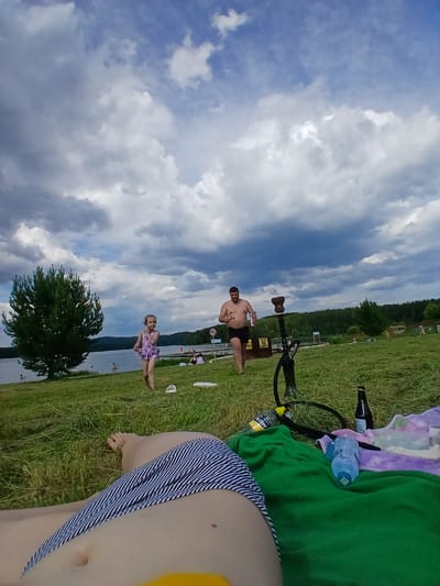 In Revda, Russia, a group of people are enjoying a sunny day outdoors, possibly a picnic or a day at the beach. The scene is set by a body of water, likely a lake or river, with a tree-lined bank in the background. The sky is filled with dramatic clouds, suggesting a dynamic weather pattern.
Two individuals are prominently featured. A man, shirtless and wearing dark shorts, stands on the grassy bank, looking towards the camera. He appears relaxed and is holding a bottle. Nearby, a young girl in a patterned swimsuit is sitting on the grass, also looking towards the camera with a gentle smile, indicating a pleasant and possibly joyful atmosphere.
Other objects scattered on the grass include a hookah, bottles of water and what appears to be alcohol, and towels. These items suggest a leisurely outing where relaxation and enjoyment are prioritized. The presence of multiple people in the background, some near the water, further reinforces the idea of a social gathering or a public recreational area.
The time of day appears to be afternoon, given the lighting and the cloud formations. The weather is partly cloudy, with patches of blue sky visible amidst the heavier clouds. The overall impression is one of a relaxed and enjoyable day spent outdoors by the water. No visible text is discernible in the provided image crops.