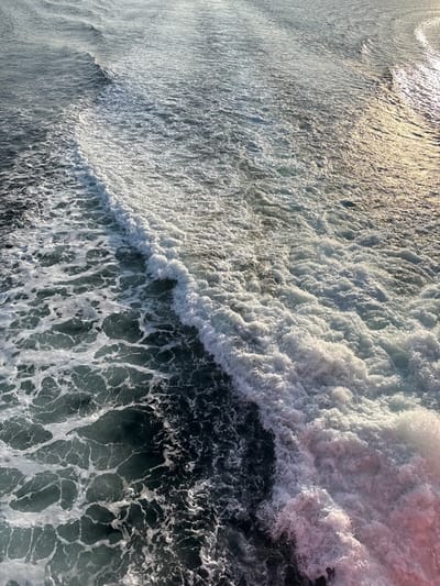 The image displays a turbulent body of water, identified as a vessel's wake, located off the coast of Palaiopoli, Greece. The wake forms a diagonal division across the frame, extending from the lower left to the upper right. On the left side, the water is dark teal with a scattering of white, interconnected foam patterns. To the right, a thick, prominent band of intensely white, frothy water signifies active churning, indicating recent displacement by a moving object not visible in the frame. Sunlight reflects off the water's surface, creating bright, shimmering patches, particularly concentrated on the frothy white areas and in the upper right portion of the image, where the reflections appear golden. A faint pinkish discoloration is observable in the lower right corner of the water. No individuals, identifiable vessels, or fixed structures are present within the scene.