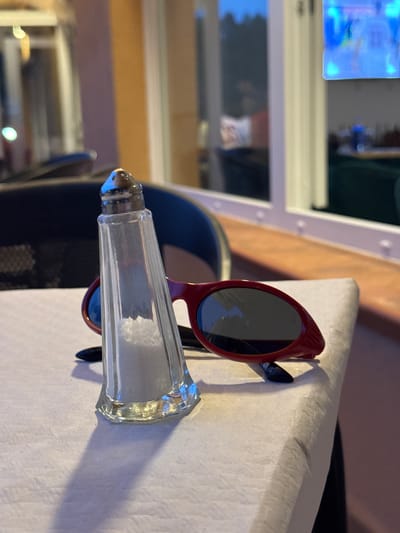 In El Escorial, Spain, on a white tablecloth, a clear glass salt shaker filled with salt is positioned in the foreground. The salt shaker has a metallic top with holes. Behind it, a pair of red-rimmed sunglasses with dark lenses lies open. The scene appears to be indoors, with a window in the background showing an out-of-focus view of trees and a television screen. The lighting suggests it is either evening or a dimly lit interior.