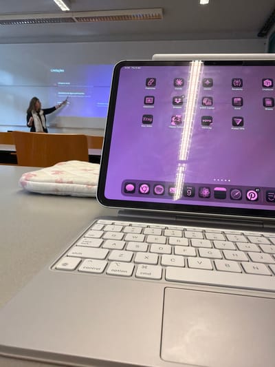 The image is taken in a classroom setting, likely in São Mamede de Infesta, Portugal, given the text in Portuguese projected on the screen ("Limitações"). The classroom has a whiteboard with an image projected onto it, showing a woman presenting information. A desk or table occupies the foreground, on which sits a closed iPad with a keyboard attached. The iPad screen displays an array of app icons with a purple background. A pillow rests on the desk in the middle of the frame. The composition of the image suggests it was taken from an elevated point of view.