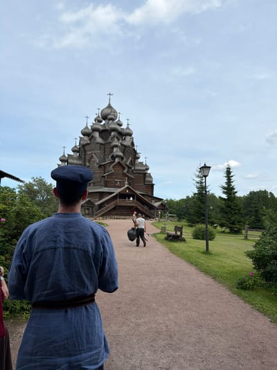 In Nevskii Parkleskhoz, Russia, a person in a blue traditional garment and cap faces away from the camera, walking on a gravel path towards a large, intricate wooden church. The church, with its numerous domes topped with crosses, is the focal point of the background. The path is lined with green grass and trees. Two other individuals are visible in the distance, walking on the same path. One carries a black trash bag, and the other is closer to the church entrance. The sky is cloudy, with patches of blue visible. The environment suggests an outdoor historical or cultural site.