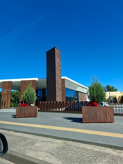 The image shows a modern building with a tall, dark brown clock tower in front. The building has a white roof and large glass windows. Two large, rectangular planters with evergreen trees and red flowers are placed on the grey paved area in front of the building. A yellow tactile paving strip is visible on the ground. In the background, there are trees and another building. The sky is clear and blue. The scene appears to be the Jurbarkas Railway Station in Jurbarkas, Lithuania.