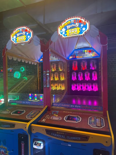 Two 'Down the Clown' arcade game machines from the 'Carnival Classics' series are depicted indoors in Saint-André-lez-Lille, France. Each machine features a red and white striped tent-like canopy with an illuminated sign displaying 'BONUS 500 TICKETS.' The machine on the left has a netted playfield with several glowing green circular targets and vertical rainbow-colored light bars at the base. The machine on the right displays three rows of stationary clown figures, illuminated in red, yellow-orange, and purple-pink, respectively from top to bottom. The control panels are brown, showing digital scores (e.g., '1105' on the right machine) and game input slots. The base of the machines is blue. Text visible on both machines states, 'RED LIT CLOWNS are Worth DOUBLE POINTS!' The environment includes a dark brick wall in the background and exposed metal ductwork on the ceiling. No individuals are visible in the scene.