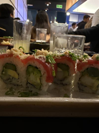 A close-up, low-angle photograph displays a plate of four sushi rolls in the immediate foreground, centered on a light-colored rectangular ceramic plate. Each roll is an uramaki style, topped with thinly sliced pink fish, green scallions, and a light-colored granular garnish. Visible fillings include slices of green avocado and a white component, possibly tempura shrimp. The background is a dimly lit restaurant interior, located in Juriquilla, Mexico. Multiple indistinct individuals are seated at tables in the midground and background. One person with long dark hair is visible from behind, and another figure wearing a dark top is partially visible on the left. Tableware, including clear drinking glasses—one containing a pale yellow liquid—and dark bowls, are visible on surrounding tables. The restaurant features dark walls, some blue accent lighting, and vertical wooden paneling, creating a modern dining environment. No specific actions or interactions are clearly discernible beyond the general dining context.