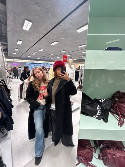 The image depicts two young adult females taking a mirror selfie within an Urban Outfitters retail store, identified by a visible shopping bag. This store is located in Westfield Mall of Scandinavia, Solna, Sweden.

The female on the left, with long light-brown hair, is wearing a long black coat over a white sweater with a red and white patterned design, light-wash blue jeans, and dark footwear. She is holding up her right hand in a peace sign gesture and has a pouting expression.

The female on the right, holding a dark-colored smartphone with a light-colored case to capture the reflection, has dark hair and is wearing a dark red fuzzy beanie hat, a long black double-breasted coat over a brown sweater, dark trousers, and black pointed-toe footwear. She has a ring on her right ring finger and white nail polish.

The background shows a large retail space with numerous clothing racks displaying garments, several white headless mannequins dressed in apparel, and other indistinct customers. A sign partially visible in the upper background reads "COLLECT & RETURNS," and a red sign with white text is also present. To the right of the subjects, a light-green multi-tiered display shelf holds a black plastic bag printed with "URBAN OUTFITTERS" on its middle tier and multiple maroon bags with fringe details on its lower tier. The ceiling is grey and textured, featuring multiple recessed rectangular lighting fixtures. The flooring is a light, speckled surface.