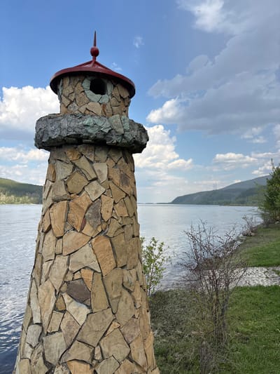 A stone lighthouse stands on the bank of the Yenisey River in the city of Овсянка, Russia. The lighthouse is built of rough, irregularly shaped tan and brown stones and is capped with a red roof and a gray stone crown. The sky is blue with scattered white clouds. The river is calm and reflects the sky. Green grass and trees line the riverbank.