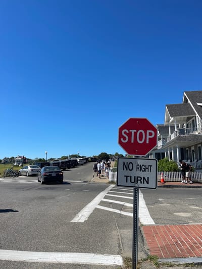 The image depicts a street scene in Oak Bluffs, Massachusetts, USA, under a clear blue sky. In the foreground, a red stop sign with white lettering is mounted on a metal pole, accompanied by a white rectangular sign below it that reads "NO RIGHT TURN." White road markings delineate a crosswalk. To the right of the signs, a white picket fence runs along the edge of a brick sidewalk and a building with grey siding, white trim, and multiple windows. Several people are visible on the sidewalk and in the street.
Further down the street, a line of parked cars stretches into the distance, with more buildings visible in the background. Several bicycles are parked on the left side of the street. The environment appears to be a sunny day in a coastal town, possibly a tourist area, given the presence of people and the architecture.