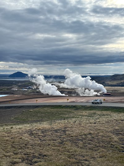 This wide-angle landscape photograph captures a scene in Reykjahlíð, Iceland, featuring active geothermal vents and a vast, natural environment. In the mid-ground, large plumes of white steam rise prominently from a geothermal facility or natural hot springs, dispersing into the cloudy sky. Beyond this geothermal activity, a calm body of water, likely a lake or fjord, stretches towards distant, rugged mountains under an expansive, partially overcast sky with varying shades of grey and brighter patches.

A paved road runs across the mid-ground of the image, with a narrower dirt shoulder alongside it. On the right side of the dirt shoulder, a white SUV is parked, facing away from the viewer. Further along the road, near the steam source, a dark brown, rectangular building or container is visible. Yellow and black chevron signs indicating a curve or hazard are intermittently placed along the roadside.

The immediate surroundings of the road and the lower slopes of the distant hills are covered in sparse, dry, brown and green vegetation, characteristic of volcanic and high-latitude landscapes. The lighting suggests it is daytime, possibly late morning or afternoon, with diffused light from the cloudy conditions. No discernible text is visible in the image. The scene conveys a sense of raw natural power and the unique geological features of Iceland.