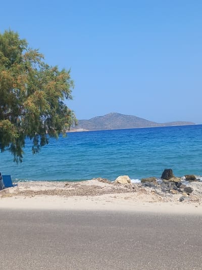 The image depicts a serene coastal scene in Zipari, Greece, under a clear blue sky. In the foreground, a gray asphalt road runs horizontally across the lower part of the frame. Beyond the road lies a sandy beach, scattered with dry seaweed and small rocks. To the left, a single green tree with a dense canopy extends into the frame, its branches partially obscuring the view of the sea. Two blue lounge chairs are visible on the beach, suggesting a place for relaxation. The sea itself is a vibrant turquoise, with gentle waves lapping at the shore. In the distance, a range of low, undulating hills forms the horizon. The overall atmosphere is peaceful and idyllic, characteristic of a Mediterranean summer day.
