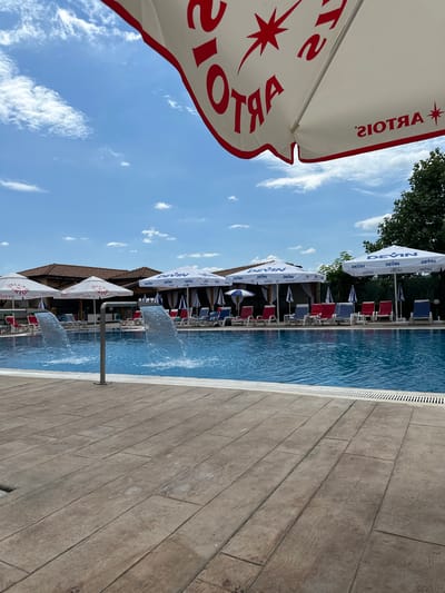 The image depicts an outdoor swimming pool area on a sunny day. The pool itself is a vibrant blue, with water gently flowing from two modern fountain structures. Around the pool, there are numerous lounge chairs in red and blue, arranged beneath several large white umbrellas. Some of these umbrellas are branded with the logo "DEVIN," while others display the "STELLA + ARTOIS" logo. In the background, a covered seating area with wooden beams and a tiled roof is visible, suggesting a resort or restaurant setting. The ground around the pool is paved with a wooden-patterned concrete. The sky above is a clear blue, dotted with fluffy white clouds. The overall scene conveys a sense of relaxation and leisure.