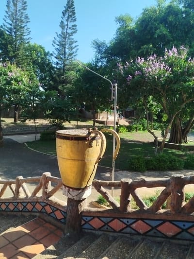 The image shows a decorative, oversized, yellow drum with handles, mounted on a decorative concrete post. The drum sits atop a stairway with an ornately painted concrete railing in a public space in An Phu Ward, Vietnam. The stairway is lined with orange-colored tiles, and the railing is painted with blue and orange diamond patterns. The backdrop includes green trees, some with purple flowers, a grassy area, and a light pole. The sky is blue and clear.