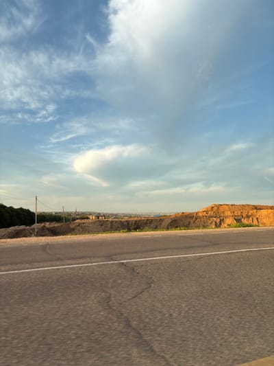 The image shows a wide asphalt road with a white line marking the lanes. To the right of the road, there is a large pile of dirt and sand, with distinct layers. Beyond the dirt pile, a line of trees forms the background on the left, and a distant town with buildings can be seen in the center. The sky is blue with scattered clouds. The overall scene depicts an area undergoing construction or excavation, likely in a rural or suburban setting. The specific location is Plodopitomnik, Russia. There are no people or animals visible in the image. The lighting suggests it is either early morning or late afternoon.
