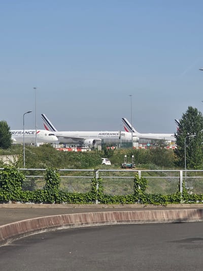 The image, taken in the city of Mitry-Mory, France, depicts an airport tarmac on a clear day. Two large Air France airplanes are parked side-by-side, with their tails clearly visible. The airplane on the left has "AIRFRANCE" written in large letters on its fuselage. Both aircraft appear to be passenger jets. In the foreground, a chain-link fence covered in ivy runs horizontally across the frame, separating the viewer from the airport area. Beyond the fence, there is dense green foliage and several trees. A white car is parked to the left of the aircraft. In the background, tall light poles are visible, characteristic of an airport environment. The ground in the immediate foreground is asphalt with a concrete curb.