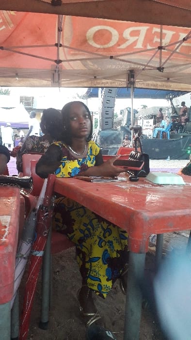 The scene depicts an outdoor gathering in Agukwu Nri, Nigeria, under a large, orange-colored canopy tent. The tent's underside displays reversed text, including "ZERO" (likely "HERO" beer) and "LAGER," along with a warning "Not for sale to persons under the Age of 18."
In the foreground, a young female individual, dressed in a yellow and blue floral patterned Ankara fabric dress and a silver-colored necklace, is seated at a red plastic table. Her dark hair is styled, and she wears silver-colored bangles on her left wrist and dark sandals with metallic straps. She holds a dark-colored smartphone in her right hand and is looking towards the viewer.
On the table in front of her rests a black purse, a smaller black object, and a light-colored rectangular item. To her left, an umbrella with red and grey sections leans against a red table leg. Behind her, another individual is partially visible, seated at a red plastic chair. The ground beneath the tables appears to be dirt.
In the mid-ground, a stage is set up, featuring large black speaker stacks on the left. Several individuals are visible on and around the stage, some appearing to be musicians or performers. One person in a light blue garment is seated on a chair on the stage. Additional white tents or structures are present in the distant left background.