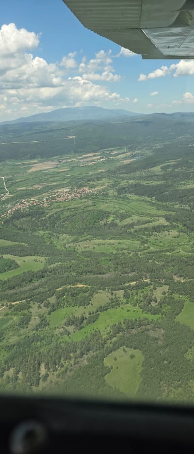 The image, taken from an elevated position, possibly from an aircraft, displays a landscape near the city of Dragushinovo, Bulgaria. A mix of forested areas, open fields, and scattered settlements characterize the terrain. In the central region, a village with clustered buildings is visible, interspersed with cultivated farmlands. Forests dominate the upper and lower portions of the image, exhibiting varied shades of green. The sky is blue with scattered white clouds, and the aircraft's wing can be seen on the left. The presence of valleys and rolling hills suggests a moderately mountainous region.