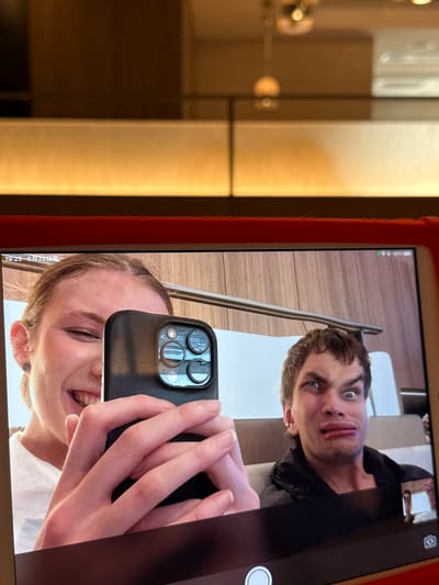 This image shows a tablet screen displaying a video call between two individuals. On the left, a young woman with blonde hair pulled back smiles widely and holds up a black smartphone, partially obscuring her face. Her right hand is visible, holding the phone which has a triple-lens camera system. On the right side of the screen, a young man with brown hair makes a distorted facial expression, his eyes wide and his mouth puckered. He is wearing a black jacket and appears to be sitting on a seat. A small thumbnail image of the woman is visible in the bottom right corner of the man's frame. The tablet screen shows a date and time stamp at the top left that reads "19:25 6月22日周一," indicating it is 7:25 PM on Monday, June 22nd. The battery level indicator at the top right shows 100%. The background on the tablet screen appears to be a wooden-paneled wall. The tablet itself is housed in a red protective case. The background of the original image shows a blurred indoor setting with warm lighting. While the image contains a date and time which could potentially be associated with Shanghai, China, there is no visual information within the image itself to confirm this location.