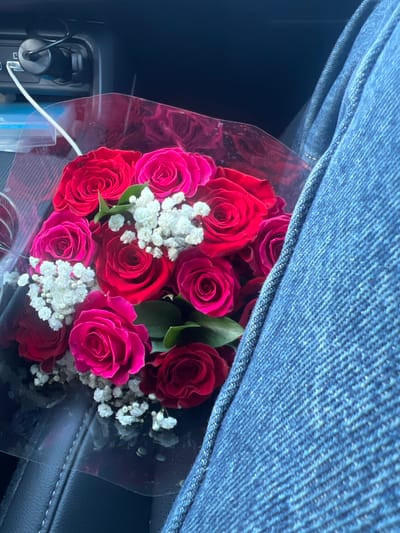 A bouquet of red and pink roses is wrapped in clear plastic. Small white flowers are interspersed among the roses. The bouquet rests on a dark surface, possibly leather, with a section of denim fabric draped over the top. The scene is close-up, capturing the vibrant colors and textures of the flowers and surrounding materials. A charging cable is visible on the left.