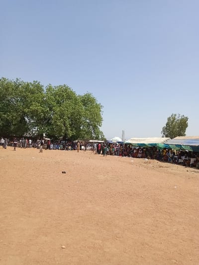 A large gathering of people is seated under white and green canopies in a wide, sandy field. The field is mostly bare, but a few trees are visible in the background. The sky is clear and blue. The scene appears to be a large public event, possibly a festival or a political rally. The location is likely Zum, Nigeria, based on the coordinates provided. The people are seated in rows, facing towards a stage that is not visible in the image.