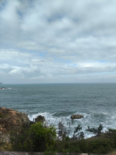 An overhead photograph depicts a rugged coastal scene in Tuy Phong 4, Vietnam, under an overcast sky. The upper two-thirds of the frame are dominated by a dense, light grey cloud cover with minimal patches of blue sky visible. The mid-ground features a vast expanse of dark teal-green ocean with visible ripples and waves. In the lower left, a rocky coastline with reddish-brown and grey geological formations descends towards the water, where white sea foam is present from breaking waves. Another isolated dark rock is situated in the turbulent, white-capped water in the lower right. The immediate foreground consists of dense green and sparse, drier vegetation, including bushes and small trees. A distant, dark landmass or mountain range is faintly visible on the horizon in the far left, extending into the water, alongside smaller dark rock formations. Several minute, dark specks, possibly boats or buoys, are scattered across the mid-ocean. No individuals, explicit actions, or direct interactions are observable. The lighting is diffuse, indicating an overcast day.