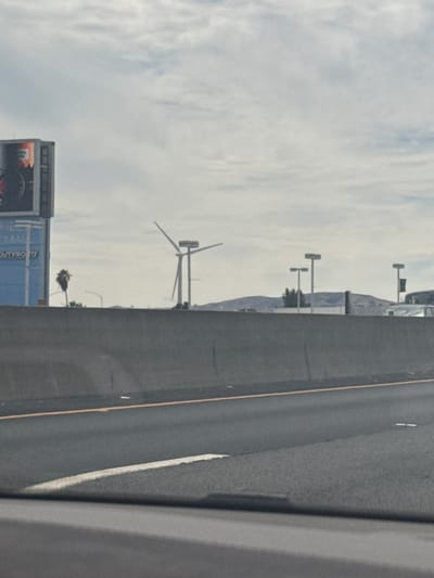 The image depicts a highway scene with a concrete barrier in the foreground. In the background, a billboard is visible on the left, and a wind turbine stands to the right of the billboard. Further in the distance, rolling hills and several streetlights can be seen. The sky is overcast with clouds. The location is the city of Fairfield, United States.