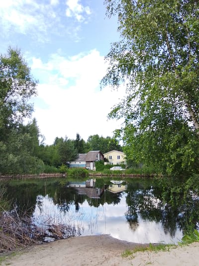 This media file depicts a serene natural scene with human habitation in Snopok Novyi, Russia. The foreground shows a sandy bank leading to a still body of water, likely a lake or river, that reflects the surrounding environment. Dry reeds and grasses are visible along the water's edge.
The middle ground features a line of trees and vegetation along the opposite bank. Nestled within this greenery are two houses. The house on the left is partially obscured by trees and appears to be a single-story structure with a light-colored facade and a darker roof. To its right is a yellow, two-story house with a white roof and visible windows. A white car is parked in front of the yellow house.
The background is dominated by a lush green forest and a sky filled with scattered white clouds against a pale blue backdrop. The reflections in the water create a mirror image of the houses and the sky, adding depth and tranquility to the scene. The overall impression is one of a peaceful, rural summer day. The lighting suggests it is daytime, likely late morning or afternoon, with fair weather. No visible text is present in the image.
