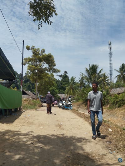 The image captures a daytime scene on a dirt road in Blangrambong, Indonesia. In the foreground, a man in a grey polo shirt and blue jeans walks away from the camera, facing right. His left foot is slightly forward, and he is wearing black sandals with a white "H" logo. To the left of the man, a dirt path is visible, with shadows cast by unseen trees.
Further down the road, another person, appearing to be a man wearing a white cap, a patterned shirt, and a sarong, walks away from the camera towards the left. He is looking over his left shoulder. Behind him, several motorbikes are parked.
On the left side of the image, a green tarp covers a structure with wooden supports. In the background, there are several houses with thatched roofs and lush vegetation, including palm trees. A tall telecommunications tower stands in the distance, to the right of the frame. The sky is partly cloudy with white, wispy clouds. The ground is covered with dry grass and scattered debris, including plastic bottles and wrappers. The overall impression is of a rural or semi-rural setting in Indonesia on a bright, sunny day.