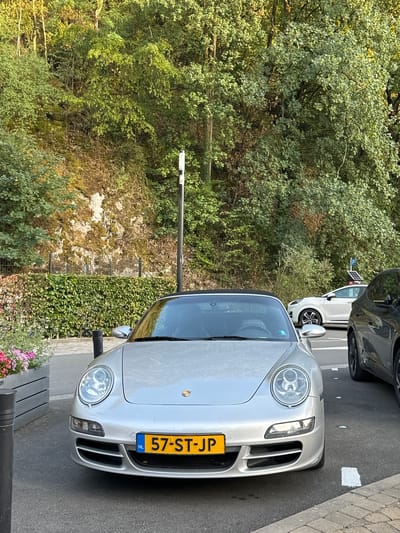 The image depicts a silver Porsche 911 parked on an asphalt surface, likely a street or parking area, in Durbuy, Belgium. The car's license plate reads "NL 57-ST-JP". To the right of the Porsche, a dark-colored SUV is partially visible, also parked. In the background, a white car is parked further down the street, and a blue parking sign with a white "P" is visible. The environment is characterized by a lush, green hillside densely covered with trees and foliage, with a rocky outcrop visible in places. A neatly trimmed green hedge runs along the base of the hillside, separating it from the paved area. In the foreground, a planter with pink and white flowers is situated to the left of the Porsche. The lighting suggests it is daytime, with sunlight illuminating the scene and creating shadows. The scene is static, with no visible people or activity.